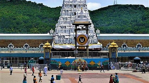 A wide shot of the main entrance (Maha Dwaram) to the Tirumala Venkateswara Temple in Tirupati, India. The tall, multi-tiered white and silver gopuram stands prominently against the sky. A central mandapam (pavilion) with a large 'U' shaped emblem is visible just below the gopuram. A large, open courtyard or plaza is filled with devotees. Green hills covered in foliage rise behind the complex.