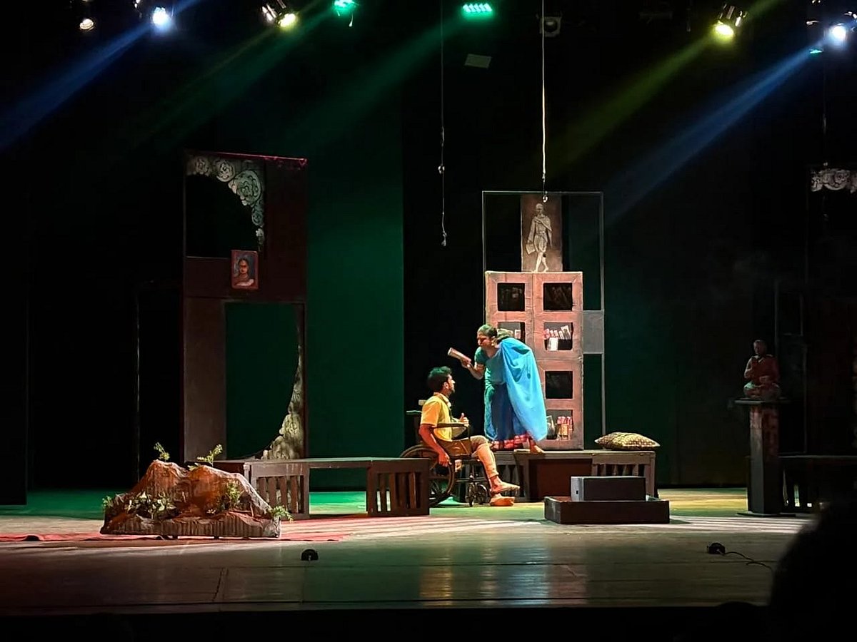 A scene from 'Bhagavantana Marana' : A male and female actor on stage in conversation. The woman is standing on a bench and looking down on the man who is sitting in a wheelchair