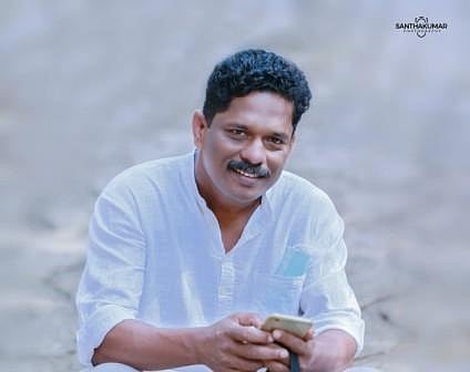Dr Sam Kutty in a white shirt, seated in a blurred background, holding a mobile phone and smiling at the camera