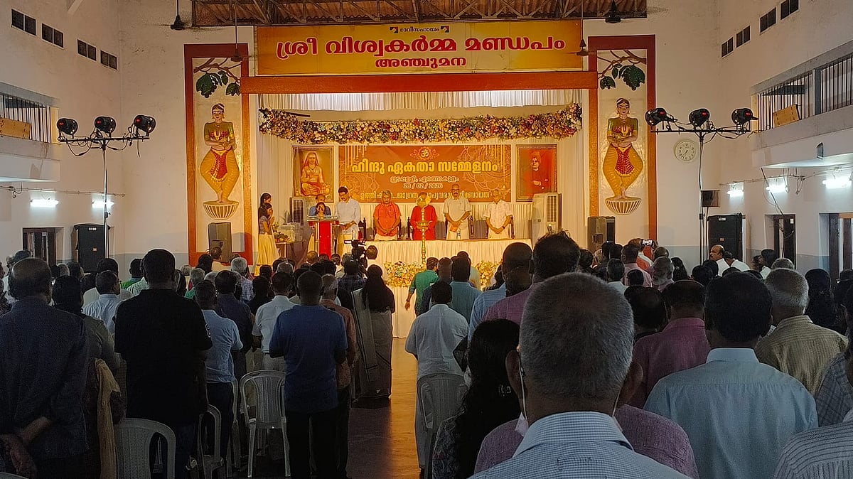 Participants gather inside a hall during a Hindu Ekta Sammelanam, facing a decorated stage where speakers stand near a traditional lamp.