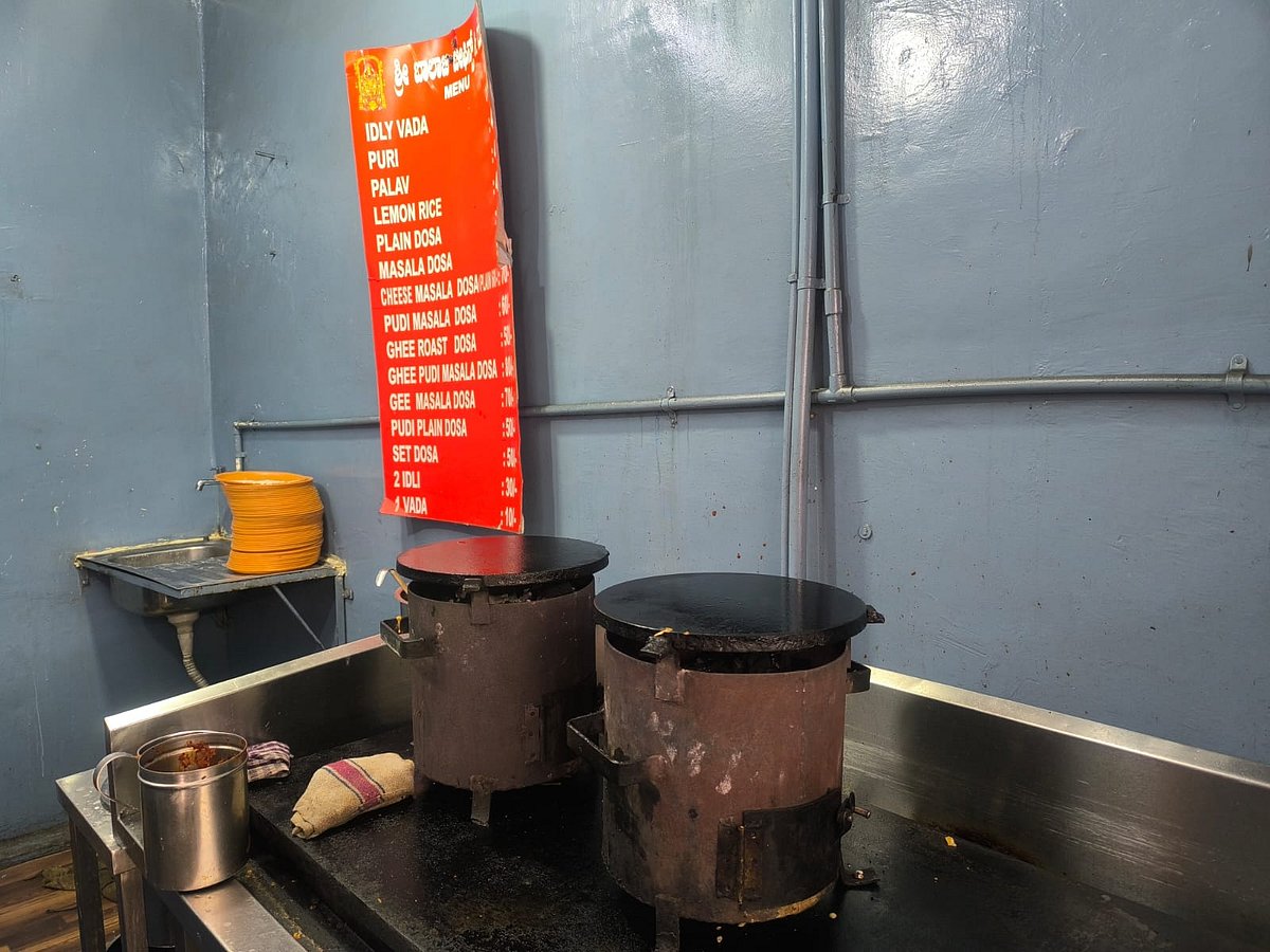 The interior of a small Indian tiffin center featuring a stainless steel preparation counter. On the counter sit two large, cylindrical metal stoves with flat, black circular griddles (tawas) on top, likely used for making dosa. To the left, there is a small metal container, a cloth, and a sink with a stack of orange plates. Mounted on the light blue wall behind the counter is a bright red menu board listing various South Indian breakfast items like Idly Vada, Puri, and various types of Dosa, with text in both Kannada and English. Translucent grey conduits run vertically along the wall.