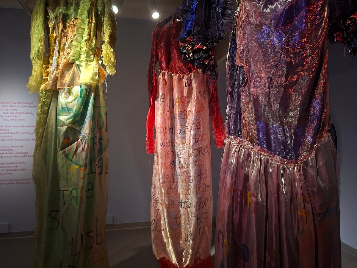 Three long, translucent garments in different colours hang side by side in a gallery space, with handwritten text visible on the fabric.