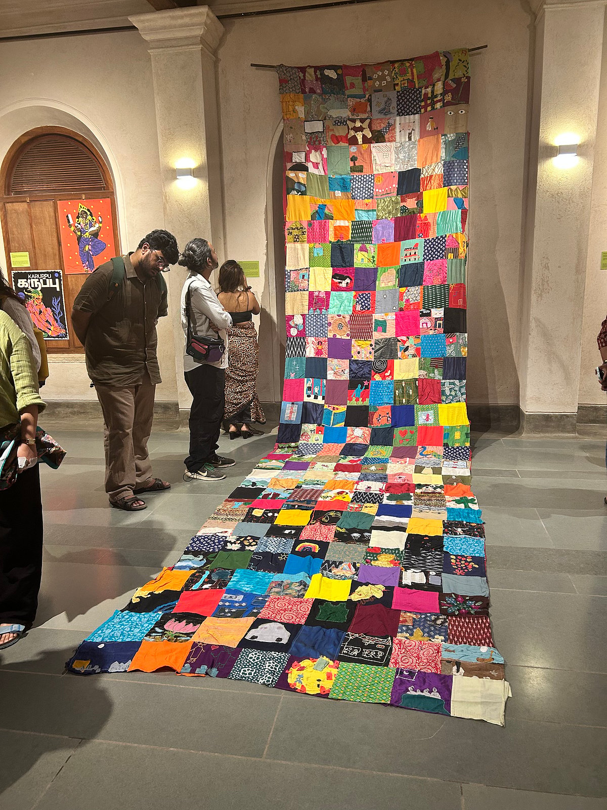 Visitors view a large, colourful patchwork textile installation made of stitched fabric squares, displayed vertically and spread across the floor in an indoor exhibition space.
