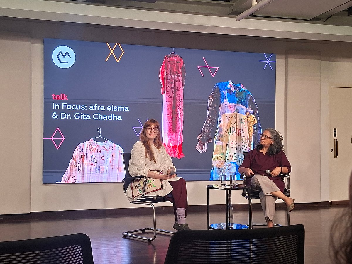 Two speakers seated on a stage during a discussion, with a screen behind them displaying images of textile garments.