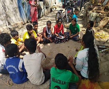 A group of children sit in a circle on a narrow street, engaged in a discussion, with modest homes and everyday street activity visible around them.
