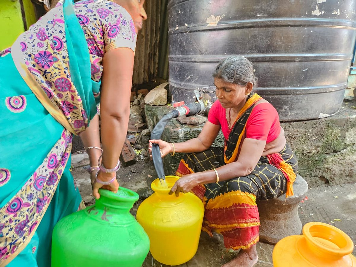 Two women in colorful saris fill plastic water containers from a pipe connected to a large storage tank in a modest outdoor setting, with one woman seated and guiding the flow while the other holds a container nearby.