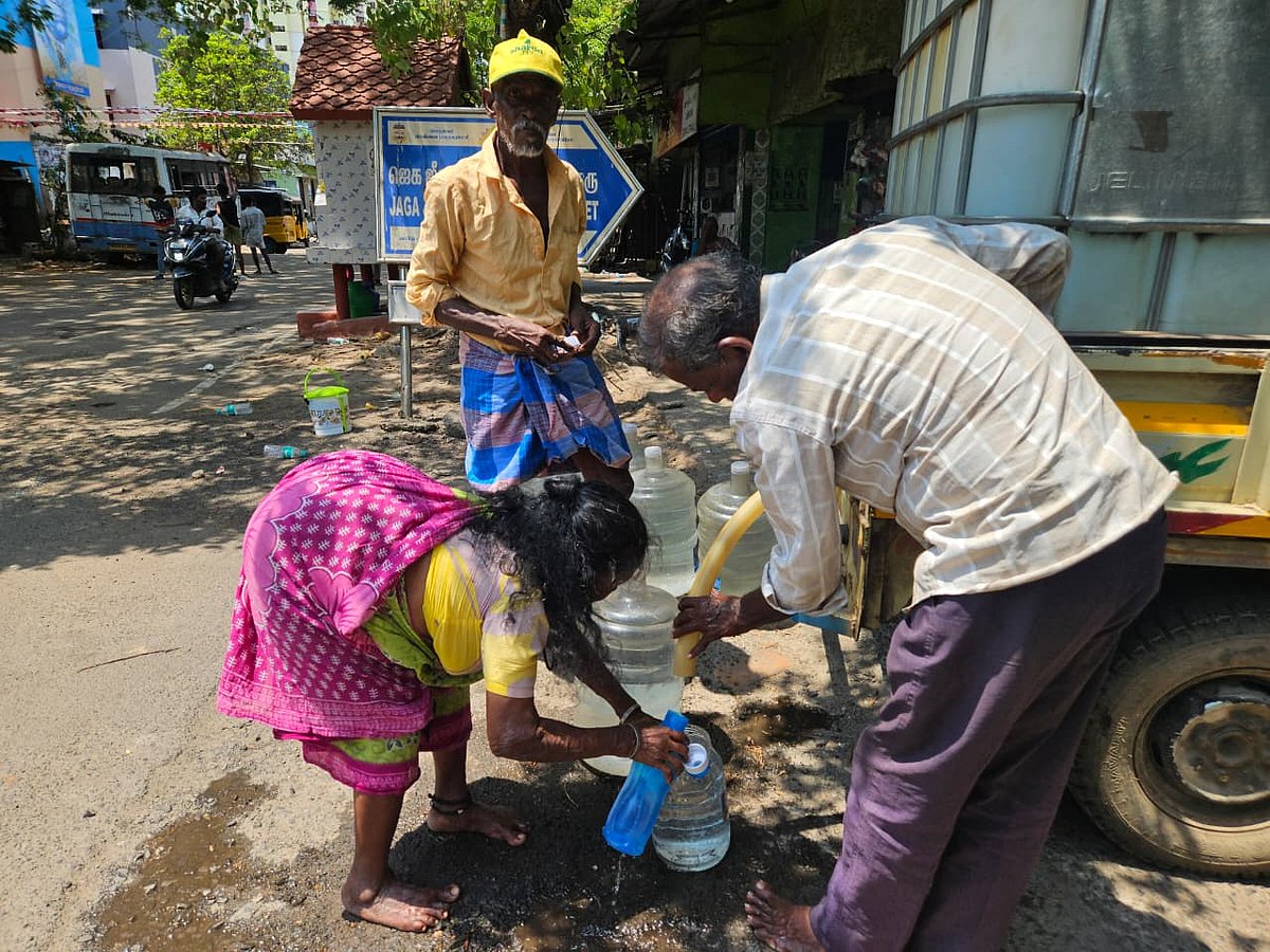 Three people fill plastic containers from a hose attached to a water tanker parked by the roadside, as others and vehicles pass in the background.