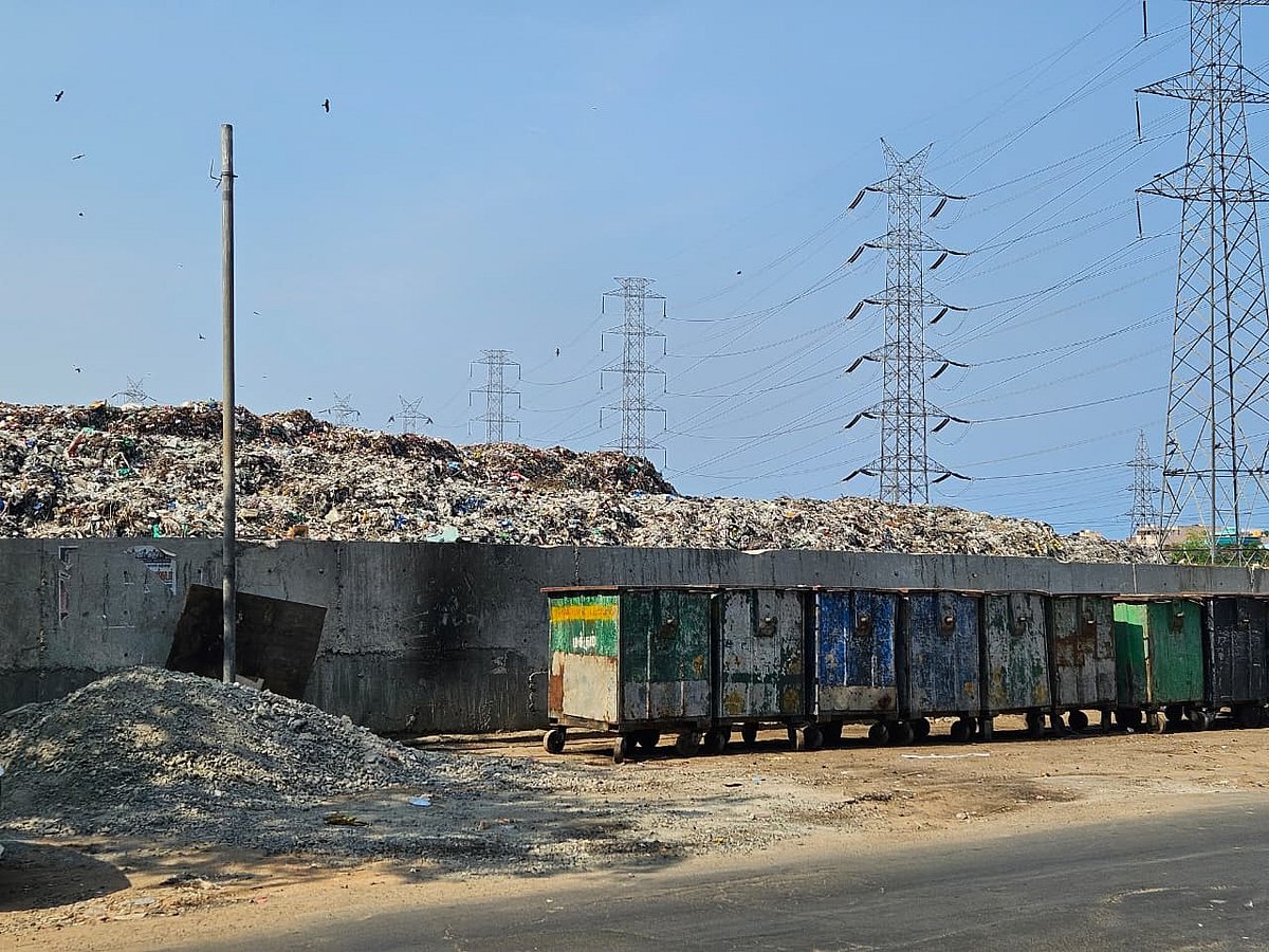 A large garbage mound towers behind a concrete wall, with several metal waste containers lined up along the roadside and electrical transmission towers visible in the background.
