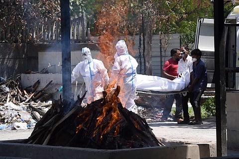 A funeral pyre burns as two men in PPE kits unload a body from the back of a vehicle with the help of two other men