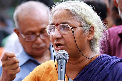A close-up photograph of Indian social activist Medha Patkar speaking into a microphone. She is an older woman with salt-and-pepper hair pulled back, wearing wire-rimmed glasses with a black strap, a red bindi, and a saffron and navy blue sari. Her expression is determined and vocal, with her right fist partially raised in a gesture of emphasis. In the blurred background, an older man with glasses is visible among a crowd.
