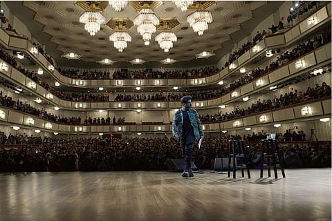 Vir Das facing the camera as he walks away from a stage in Joh F Kennedy hall with chandeliers in the background