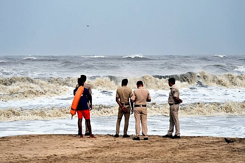 An outdoor photo shows five men, some in uniform, standing on a sandy beach looking out at rough, churning waves under a gray sky. The waves are high and foamy. Four of the men have their backs mostly to the camera, wearing light-colored uniforms that look like a police or security force. The fifth person, on the far left, wears a blue shirt and orange shorts and holds an orange life preserver, suggesting they may be a lifeguard or rescue worker. A small bird is visible near the bottom right. The image caption indicates this is related to Cyclone Biparjoy in Mumbai on June 13, 2023.