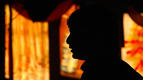 A dramatic side profile silhouette of a man with dark hair, set against a glowing, out-of-focus background of orange, red, and yellow light rectangles or windows.
