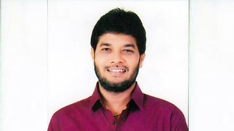 An image of a man with dark hair and a beard, wearing a purple collared shirt, smiling at the camera. The background is a plain, light color.