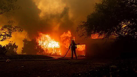 A firefighter battles an intense blaze engulfing a structure and surrounding vegetation, silhouetted against towering flames and thick smoke during a wildfire.