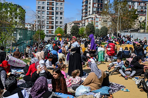 Crowds of Istanbul residents, including families with children, gather in a park during daylight after evacuating buildings due to a 6.2-magnitude earthquake, with people sitting on blankets and holding babies amid apartment buildings in the background.