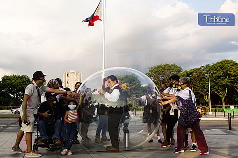 A performer stands inside a large transparent bubble as activists wearing gas masks and face masks surround him during a clean air protest at Luneta Park, Manila, with the Philippine flag in the background.