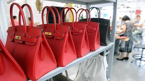 Rows of red Hermès Birkin bags are displayed on a shelf inside the Maroquinerie de la Tardoire, an Hermès workshop specializing in calfskin leather goods, in Montbron, southwestern France. Artisans can be seen working in the background.
