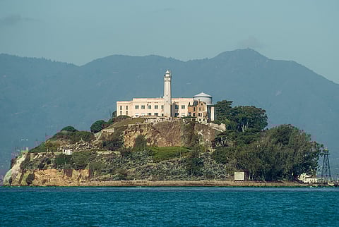 A wide view of Alcatraz Island in San Francisco Bay, showing the historic prison buildings and lighthouse surrounded by water and distant mountains.
