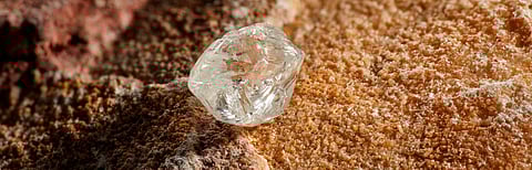 A rough natural diamond resting on a textured rock surface, symbolizing the raw beauty and origin of De Beers' mined diamonds.
