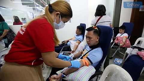 A medical worker assists a police officer donating blood during a community blood drive in Quezon City, with other participants, including a child, seen in the background.