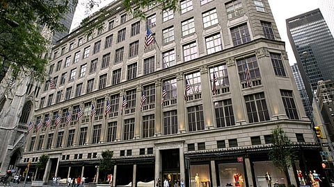 Exterior view of Saks Fifth Avenue in New York City, with American flags lining the historic building's facade. Shoppers are seen walking past display windows at street level.
