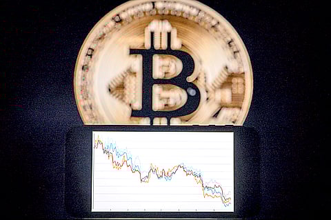 A PHONE with a downward trend in the stock market in front of a screen displaying a Bitcoin logo as the cryptocurrency’s value plummets.
