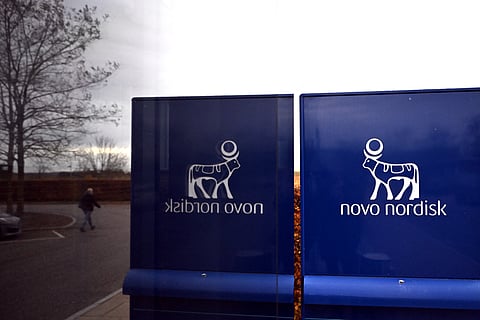 A man is reflected in a window as he walks past a sign bearing the logo of Novo Nordisk next to the company’s factory in Hillerod on November 12, 2025. On November 14, 2025 the extraordinary general meeting will take place to renew more than half of the board of directors, including the chair.